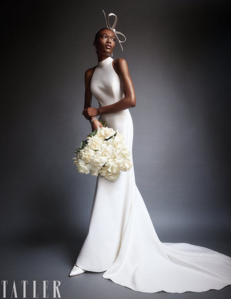 A woman in a sleek, form-fitting white gown poses gracefully against a dark background, showcasing exquisite editorial flowers. She holds a large bouquet of white roses. Her ensemble includes a unique headpiece with a looped design. The dress has a long train, and she wears pointed white shoes. "TATLER" is visible.
